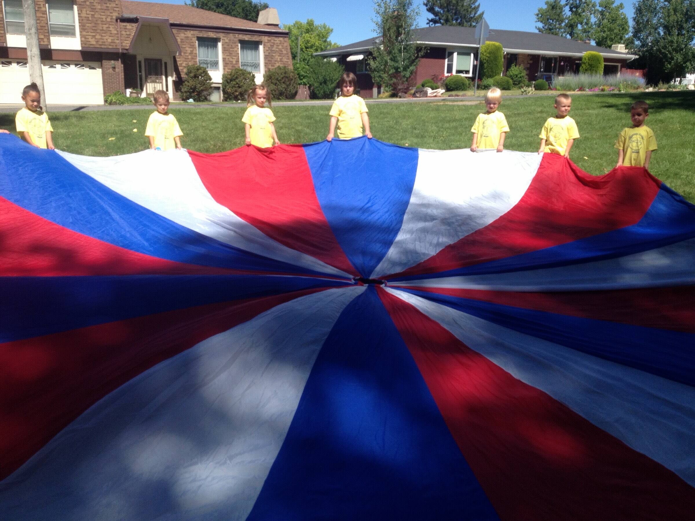 Children at Children's Corner holding a red, white and blue parachute on the lawn outside the center